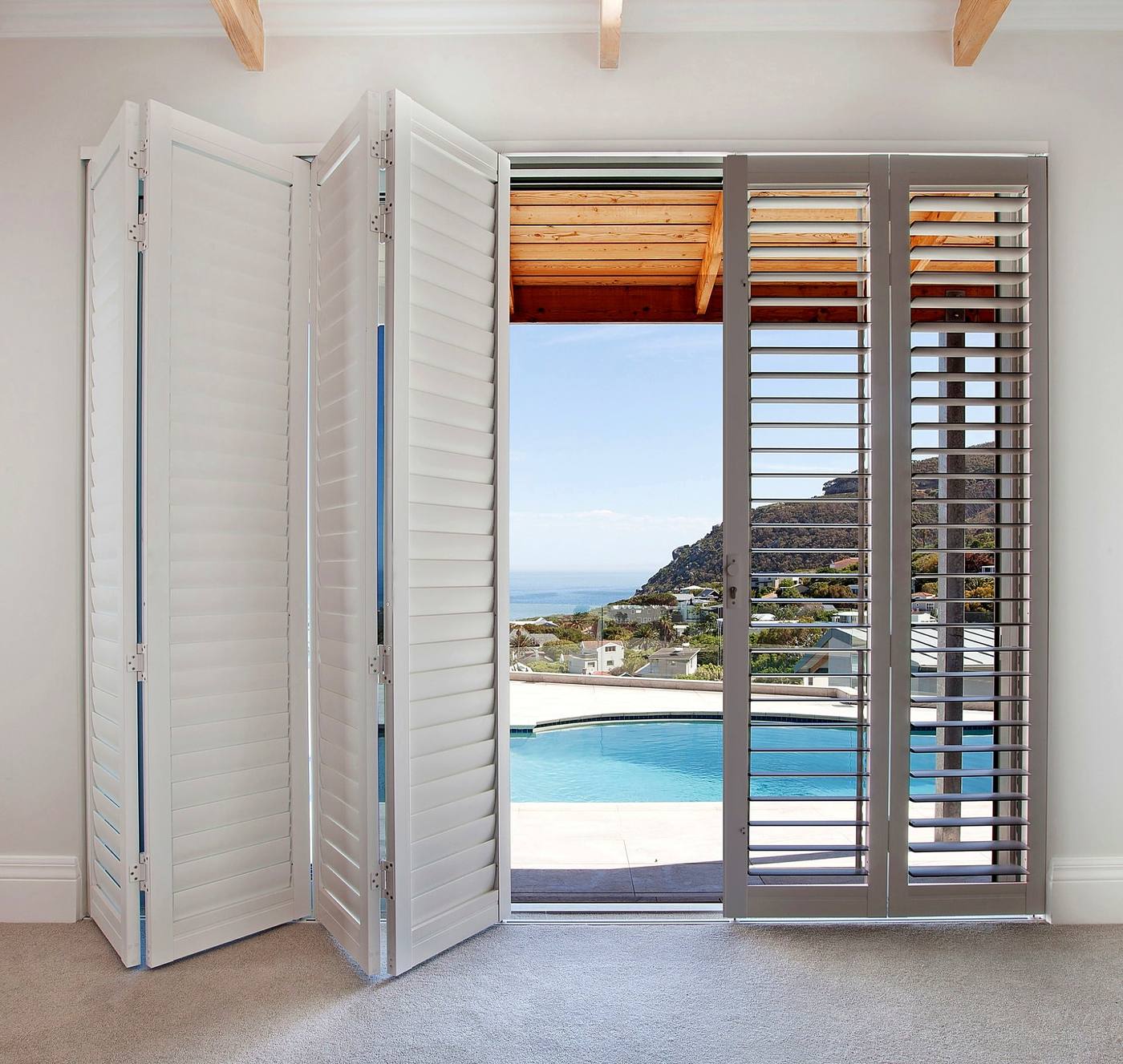 White bi-fold shutters stacked open onto a pool terrace above the coast