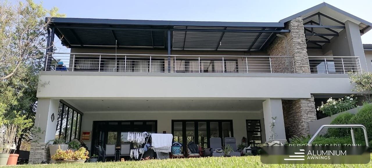 Charcoal aluminium louvre roof spanning the full-width upper-floor terrace of a double-storey Plattekloof home with a stone feature wall and stainless balustrade.